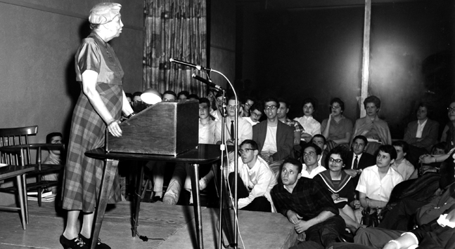 Eleanor Roosevelt stands at a small podium and mic to address students sitting in chairs and on the floor.